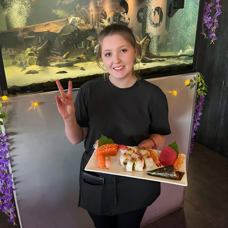 Smiling Top Sushi server holding a platter of sushi and sashimi in front of a large aquarium.