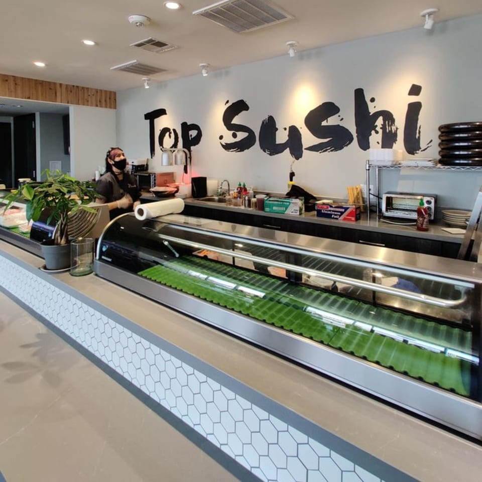 A Top Sushi employee behind the counter with a sushi display case in the foreground.