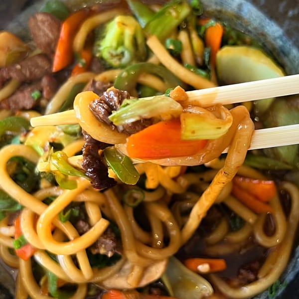 Chopsticks lifting thick udon noodles, beef, and colorful vegetables from a bowl of Top Sushi's stir-fry.