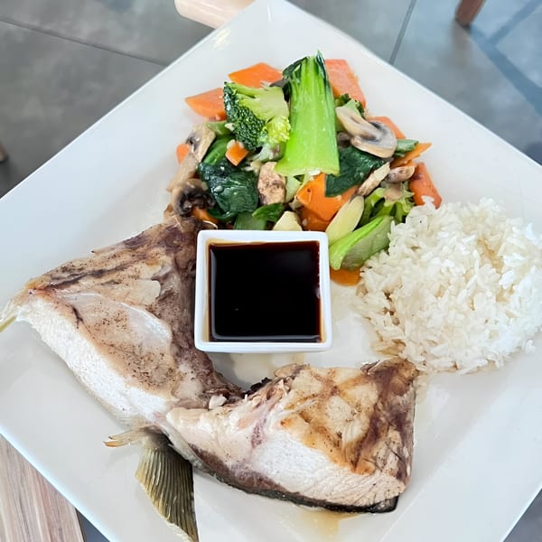 Grilled fish with rice, steamed vegetables, and a side of soy sauce on a white plate.